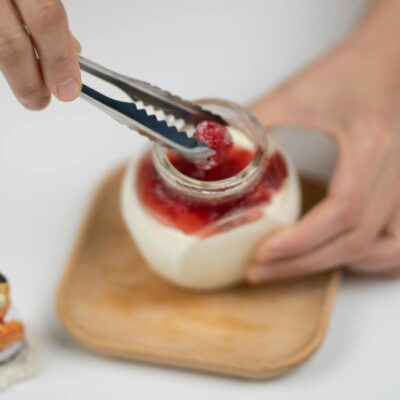 A person using tongs to place fresh raspberries on homemade yogurt dessert in a jar.