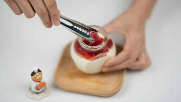 A person using tongs to place fresh raspberries on homemade yogurt dessert in a jar.