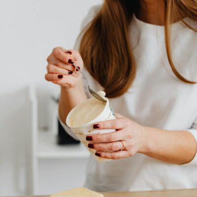 Woman with manicured nails holding yogurt container and spoon, standing by a wooden table in a kitchen.