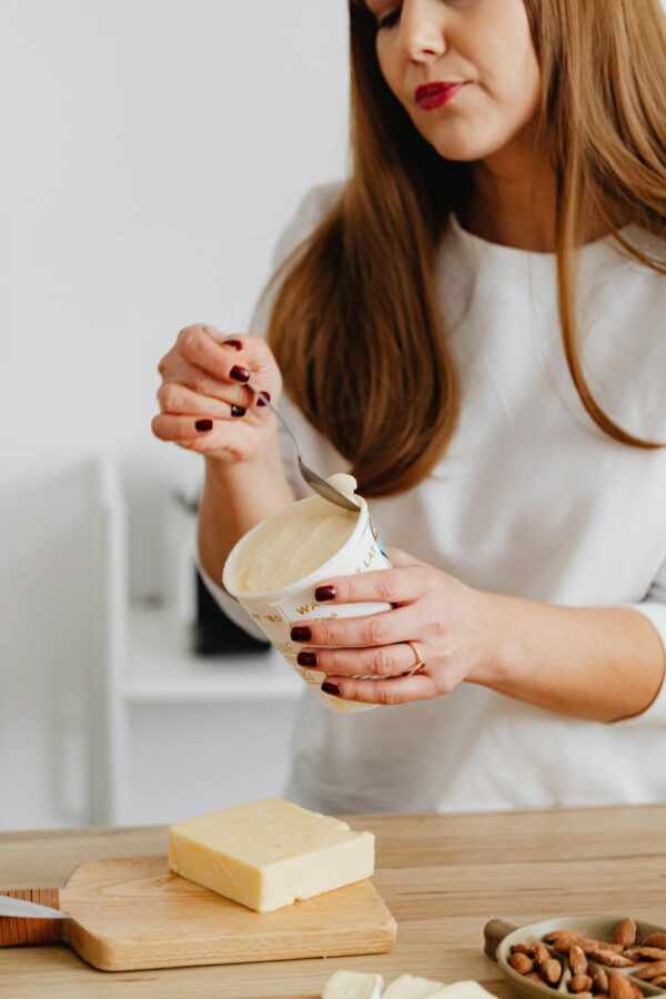 Woman with manicured nails holding yogurt container and spoon, standing by a wooden table in a kitchen.